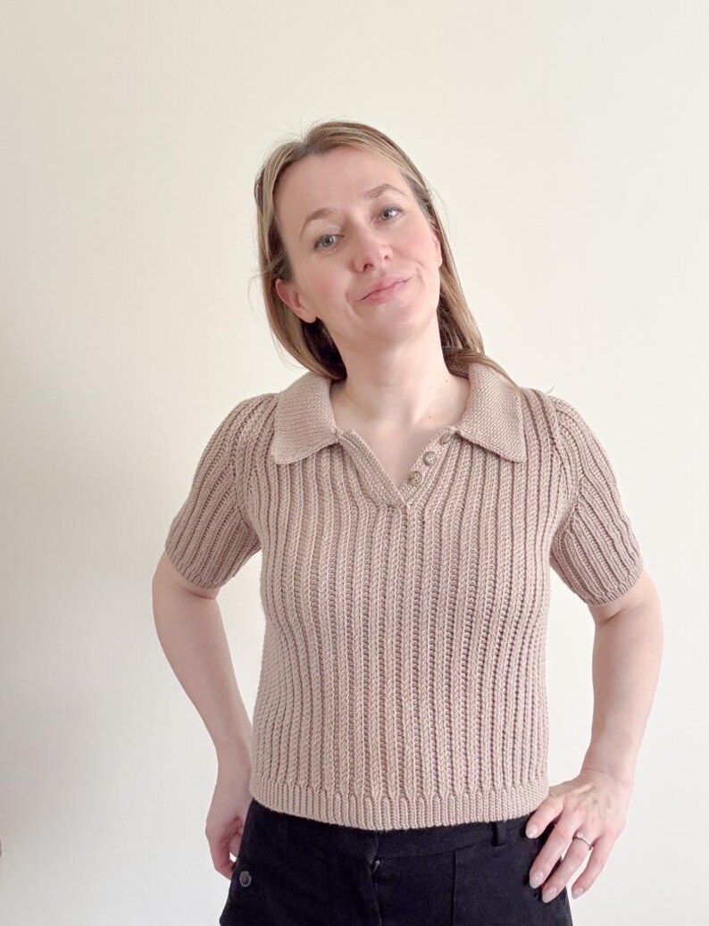 May include: A tan-colored, short-sleeved knit polo shirt with a collared neckline and three buttons. The shirt has a ribbed texture and a V-neck design. The model is wearing black pants.