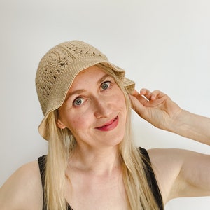 A blond woman is wearing a beige crochet bucket hat. A hat is made from granny squares with a single crochet brim. 
A woman is wearing a black tank top and there is a white background.