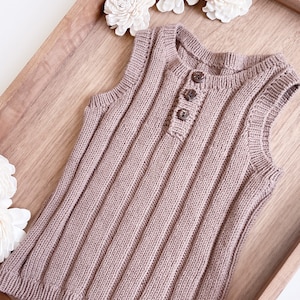 May include: A brown knitted sleeveless top with a button closure at the neck. The top is laying on a wooden tray with white decorative flowers.