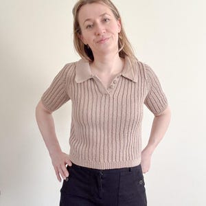 May include: A tan-colored, short-sleeved polo shirt with a ribbed knit texture. The shirt has a collared neckline and a three-button placket. The model is wearing black pants.