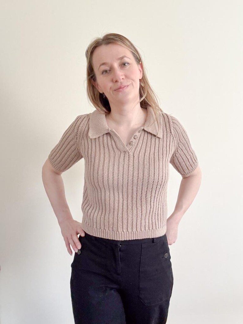 May include: A tan-colored, short-sleeved polo shirt with a ribbed knit texture. The shirt has a collared neckline and a three-button placket. The model is wearing black pants.