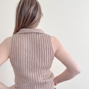 May include: A sleeveless, ribbed knit top in a neutral beige color. The top features a collared neckline and a vertical ribbing pattern. The model is wearing black pants. The top is made of a soft, comfortable material.