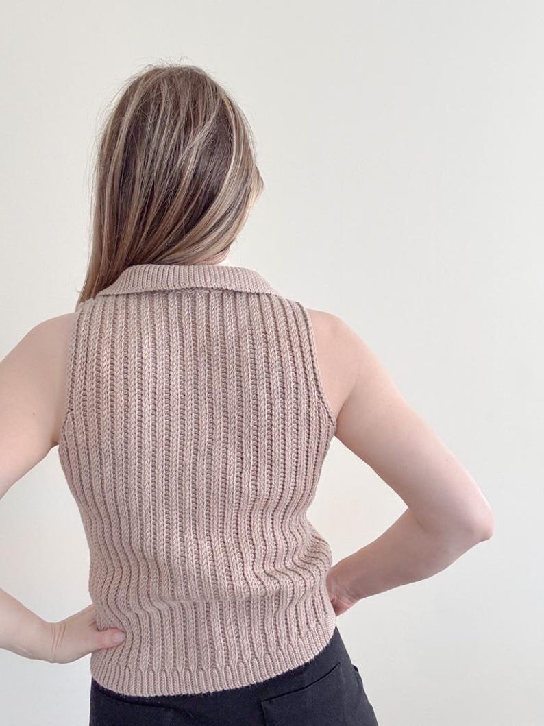 May include: A sleeveless, ribbed knit top in a neutral beige color. The top features a collared neckline and a vertical ribbing pattern. The model is wearing black pants. The top is made of a soft, comfortable material.