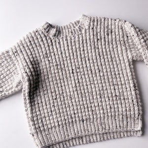 A children’s crochet sweater made from off-white worsted weight yarn (Plymouth Yarn Encore Tweed in color oatmeal 1363). Sweater is made with a textured crochet stitch looks like a chunky knit. Oversized fit, crew neck with a knit-like ribbing.