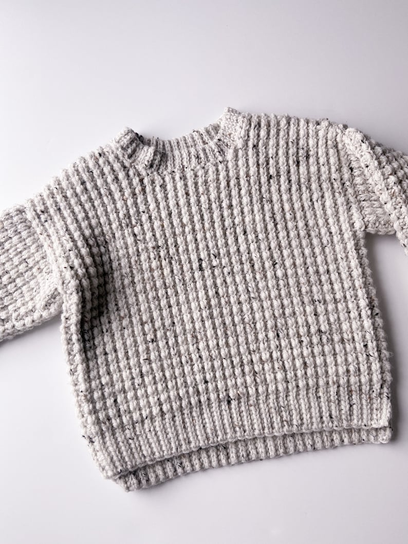 A children’s crochet sweater made from off-white worsted weight yarn (Plymouth Yarn Encore Tweed in color oatmeal 1363). Sweater is made with a textured crochet stitch looks like a chunky knit. Oversized fit, crew neck with a knit-like ribbing.