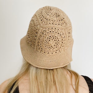 A blond woman is wearing a beige crochet bucket hat. A woman is standing facing backward to show the back of the hat. A hat is made from granny squares with a single crochet brim. A woman is wearing a black tank top and there is a white background.