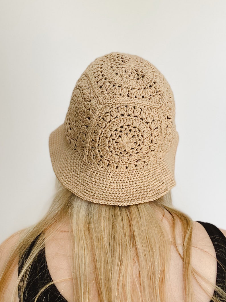 A blond woman is wearing a beige crochet bucket hat. A woman is standing facing backward to show the back of the hat. A hat is made from granny squares with a single crochet brim. A woman is wearing a black tank top and there is a white background.