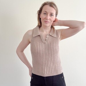 May include: A sleeveless, tan-colored knitted polo shirt with a ribbed texture. The shirt features a collared neckline with three buttons and a fitted silhouette. The model is wearing black pants.