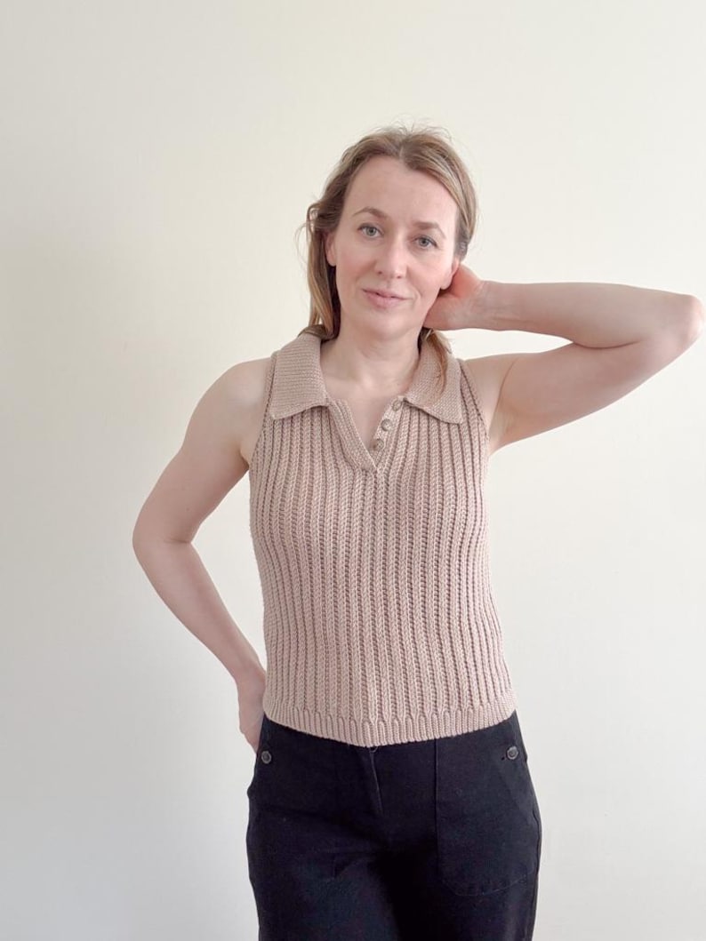 May include: A sleeveless, tan-colored knitted polo shirt with a ribbed texture. The shirt features a collared neckline with three buttons and a fitted silhouette. The model is wearing black pants.