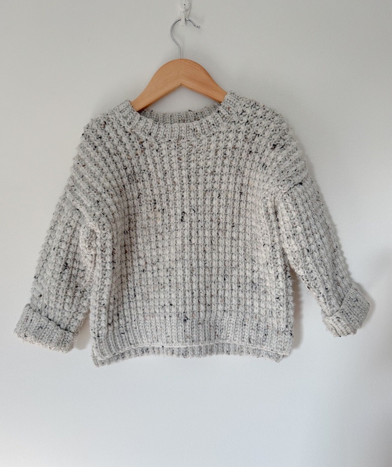 A children’s crochet sweater made from off-white worsted weight yarn (Plymouth Yarn Encore Tweed in color oatmeal 1363). Sweater is made with a textured crochet stitch looks like a chunky knit. Oversized fit, crew neck with a knit-like ribbing.