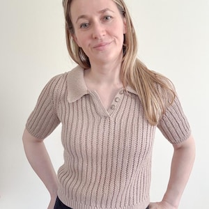 May include: A short-sleeved, tan-colored knit polo shirt with a ribbed texture. The shirt features a collared neckline and three small buttons. The model is wearing black pants.