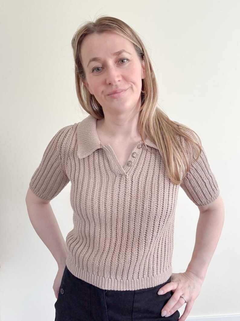 May include: A short-sleeved, tan-colored knit polo shirt with a ribbed texture. The shirt features a collared neckline and three small buttons. The model is wearing black pants.
