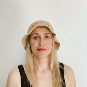 A blond woman is wearing a beige crochet bucket hat. A hat is made from granny squares with a single crochet brim. 
A woman is wearing a black tank top and there is a white background.