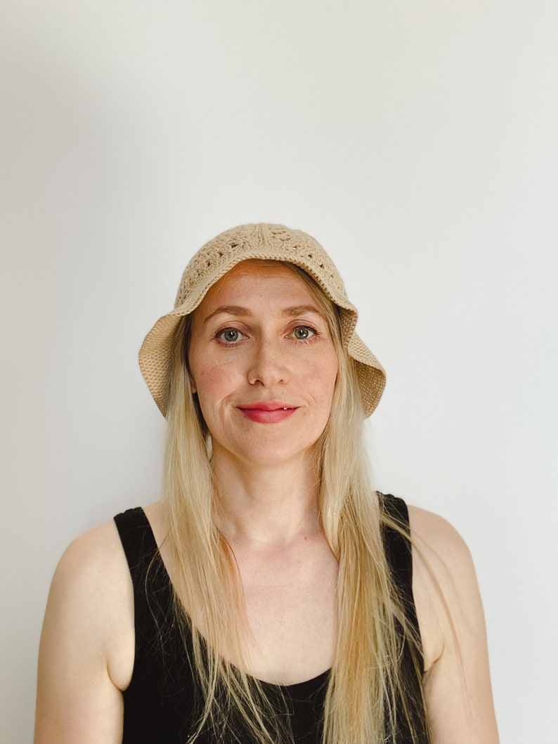 A blond woman is wearing a beige crochet bucket hat. A hat is made from granny squares with a single crochet brim. 
A woman is wearing a black tank top and there is a white background.