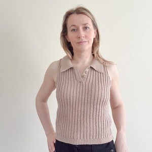 May include: A sleeveless, tan-colored knit top with a collared neckline and a V-neck. The top has a ribbed texture and two small buttons at the neckline. The person in the image is wearing black pants.