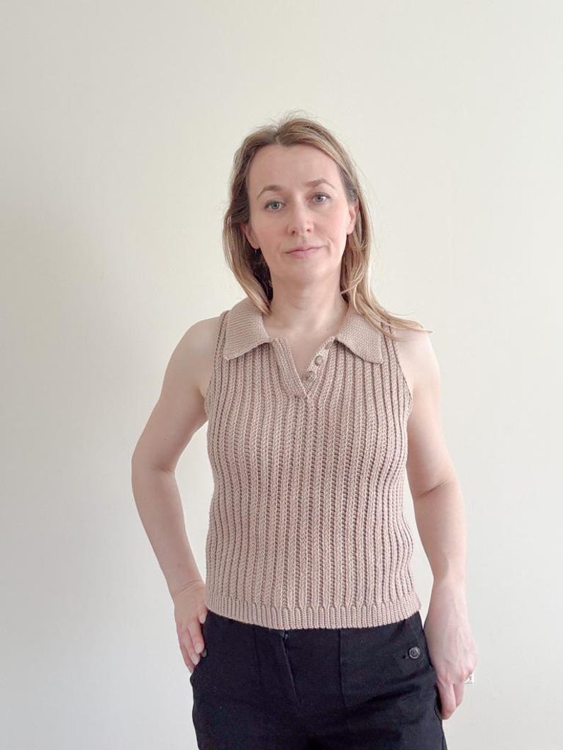 May include: A sleeveless, tan-colored knit top with a collared neckline and a V-neck. The top has a ribbed texture and two small buttons at the neckline. The person in the image is wearing black pants.