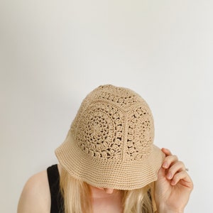 A blond woman is wearing a beige crochet bucket hat. A woman tipped her head forward to show the top of her hat. A hat is made from granny squares with a single crochet brim. 
A woman is wearing a black tank top and there is a white background.