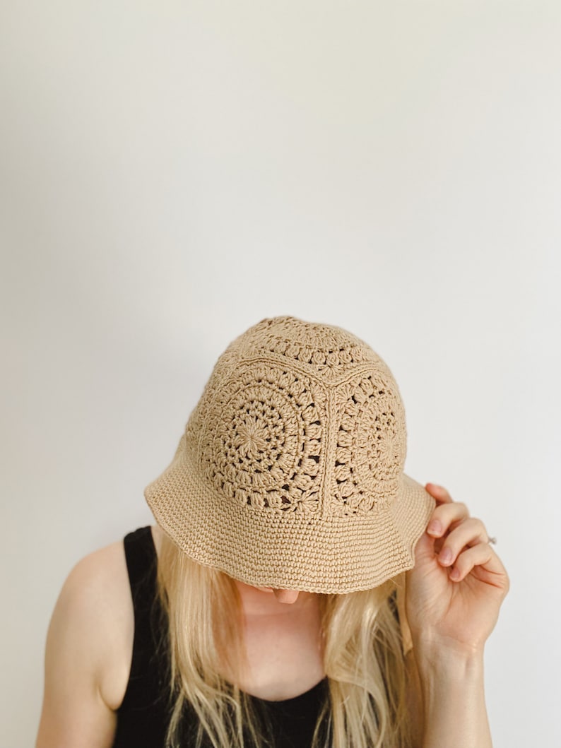 A blond woman is wearing a beige crochet bucket hat. A woman tipped her head forward to show the top of her hat. A hat is made from granny squares with a single crochet brim. 
A woman is wearing a black tank top and there is a white background.