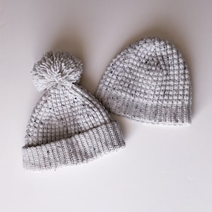 Può includere: Due berretti di lana bianca con un motivo maculato. Un berretto ha un pompon bianco soffice in cima.