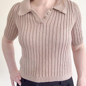 May include: A tan-colored, short-sleeved knit polo shirt with a ribbed texture. The shirt features a collared neckline and three buttons. The bottom of the shirt is visible, along with the top of black pants.