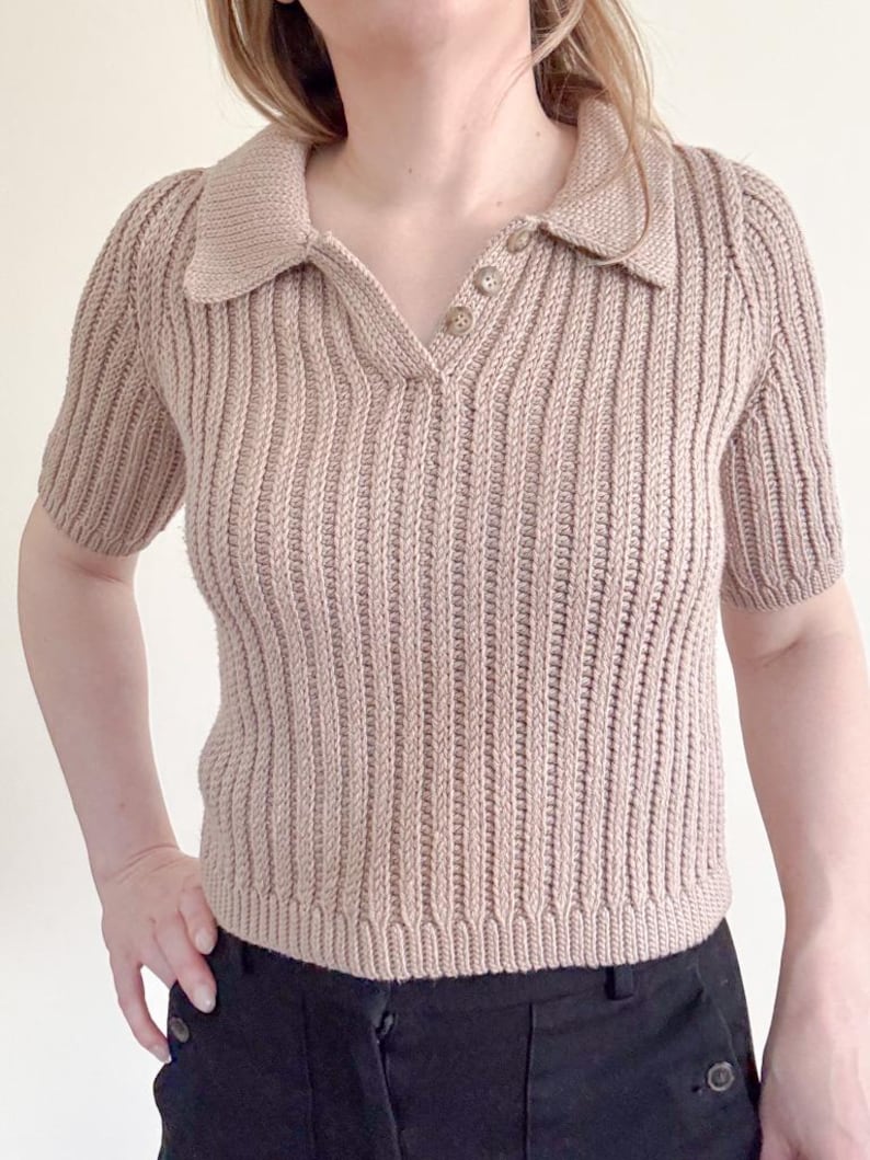 May include: A tan-colored, short-sleeved knit polo shirt with a ribbed texture. The shirt features a collared neckline and three buttons. The bottom of the shirt is visible, along with the top of black pants.