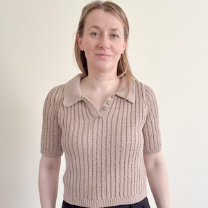 May include: A short-sleeved, beige knit polo shirt with a collared neckline and three buttons. The shirt has a ribbed texture and is paired with black pants. The overall style is casual and comfortable.