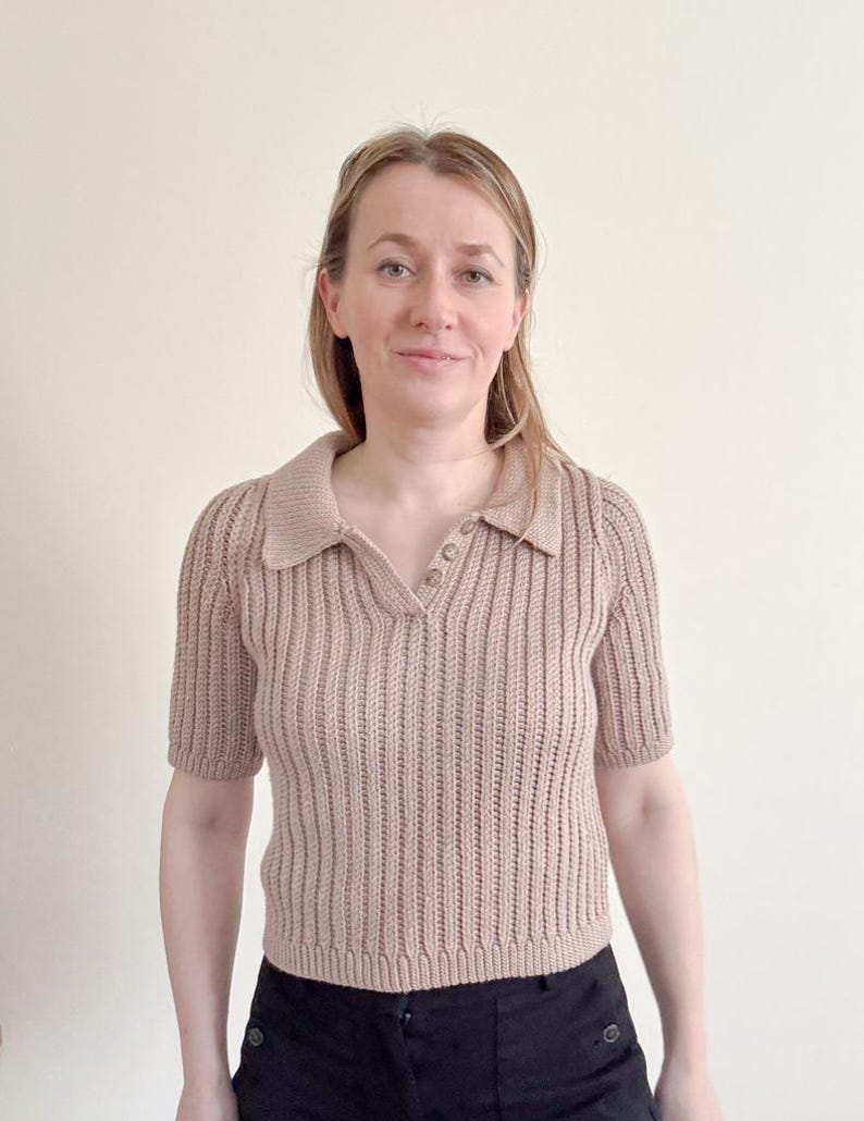 May include: A short-sleeved, beige knit polo shirt with a collared neckline and three buttons. The shirt has a ribbed texture and is paired with black pants. The overall style is casual and comfortable.