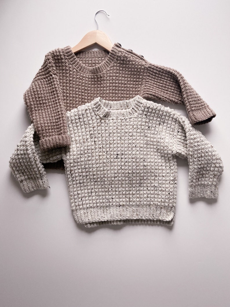 Two children’s oatmeal tweed and taupe beige crochet sweaters made with a textured crochet stitch that looks like a chunky knit. Oversized fit, crew neck with a knit-like ribbing. Taupe sweater has a shoulder button opening.