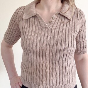 May include: A tan-colored, short-sleeved polo shirt with a ribbed knit design. The shirt features a collared neckline and three buttons. The sleeves are short and the overall style is casual and comfortable.