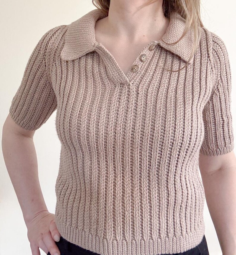 May include: A tan-colored, short-sleeved polo shirt with a ribbed knit design. The shirt features a collared neckline and three buttons. The sleeves are short and the overall style is casual and comfortable.