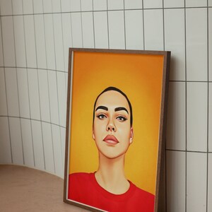 K&ouml;nnte beinhalten: Eine gerahmte Illustration einer Person mit kurzen, dunklen Haaren. Die Person blickt nach oben mit einem neutralen Ausdruck. Sie tr&auml;gt ein rotes Shirt vor einem gelben Hintergrund.