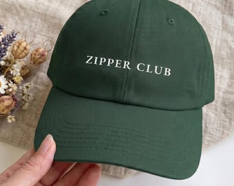 May include: A dark forest green baseball cap featuring the text ZIPPER CLUB embroidered in white on the front panel. The cap has a structured crown, a curved brim with visible stitching, and a button at the top center of the dome.