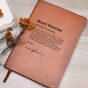 May include: A brown leather journal with the text "Heart Warrior" and a definition. The definition describes a person who has faced heart surgery with determination. The journal has a brown ribbon bookmark and the name "meghan" below the definition.