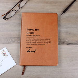 Könnte beinhalten: Ein braunes Lederjournal mit den Worten "Force for Good" und der Definition "[fors for gud] noun Eine Person, die die Welt durch nachdenkliche Handlungen und Worte besser macht, positive Veränderungen bewirkt, die weit über sich hinausreichen." Das Journal trägt auch das Wort "david" in Kursivschrift.
