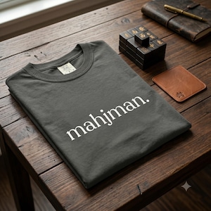 May include: A charcoal gray crew neck t-shirt featuring the white text mahjman. printed in a clean sans-serif font across the chest. The garment has a visible interior neck label and is folded neatly on a dark wooden surface.