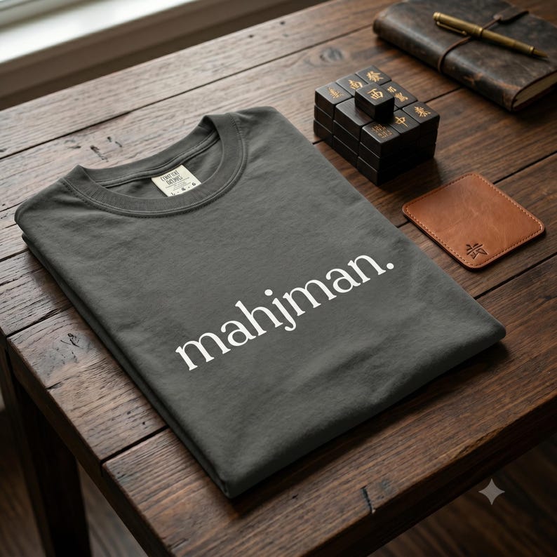 May include: A charcoal gray crew neck t-shirt featuring the white text mahjman. printed in a clean sans-serif font across the chest. The garment has a visible interior neck label and is folded neatly on a dark wooden surface.