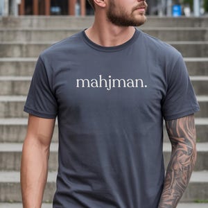 May include: A charcoal gray short-sleeve t-shirt featuring the word mahjman. printed in a white serif font across the chest. The garment has a classic crew neckline and a straight hemline, providing a clean and simple aesthetic for casual wear.
