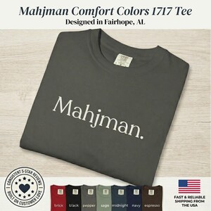 May include: A dark gray short-sleeve t-shirt featuring the word Mahjman printed in white across the chest. The shirt has a crew neckline and a Comfort Colors 1717 label. A row of seven additional t-shirts in various colors is displayed below.