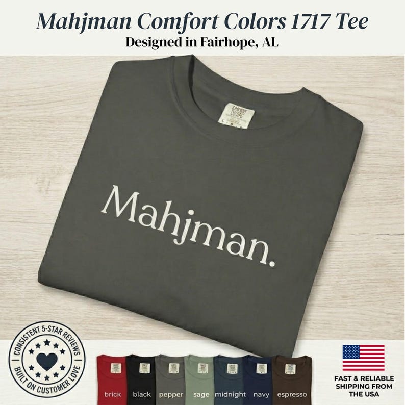 May include: A dark gray short-sleeve t-shirt featuring the word Mahjman printed in white across the chest. The shirt has a crew neckline and a Comfort Colors 1717 label. A row of seven additional t-shirts in various colors is displayed below.