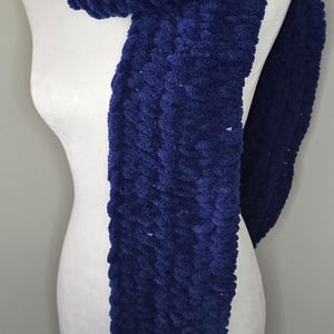 May include: A plush, navy blue scarf is draped over a white mannequin. The scarf is made of a thick, textured material, and its deep blue color is prominent. The scarf's soft texture and rich color suggest warmth and comfort, making it a cozy accessory.