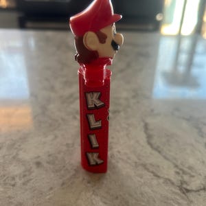 Może przedstawiać: Czerwony dozownik PEZ z postacią Mario z serii gier wideo Super Mario Bros. Dozownik ma czerwony kapelusz, brązowe włosy i czerwony korpus z nadrukiem liter "K L I K".