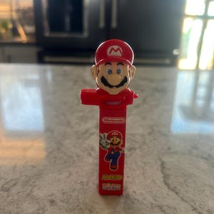 Może przedstawiać: Czerwony dozownik PEZ z postacią Mario z Nintendo. Dozownik ma czerwony kapelusz, wąsy i czerwony korpus. Opakowanie dozownika zawiera logo Nintendo i napis "Mario".