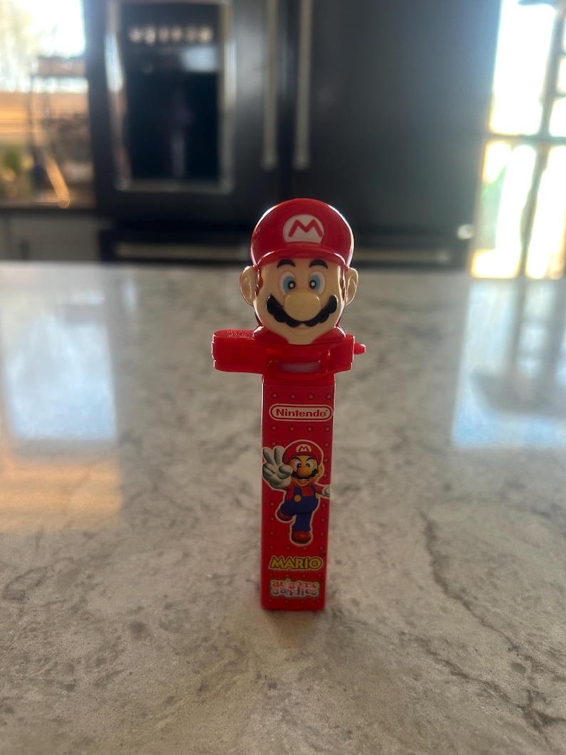 Może przedstawiać: Czerwony dozownik PEZ z postacią Mario z Nintendo. Dozownik ma czerwony kapelusz, wąsy i czerwony korpus. Opakowanie dozownika zawiera logo Nintendo i napis "Mario".