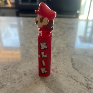 Może przedstawiać: Czerwony dozownik PEZ z postacią Mario z serii gier wideo Super Mario Bros. Dozownik ma czerwony kapelusz, brązowe włosy i wąsy. Na dozowniku wydrukowano słowo "KLIK".