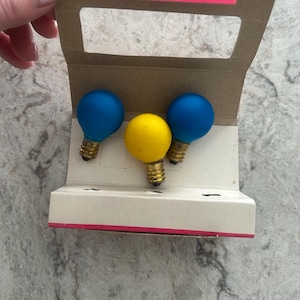 Può includere: Tre lampadine colorate in una confezione di cartone. Due lampadine blu affiancano una lampadina gialla. La confezione è beige con un accento rosa. Le lampadine hanno basi in ottone.