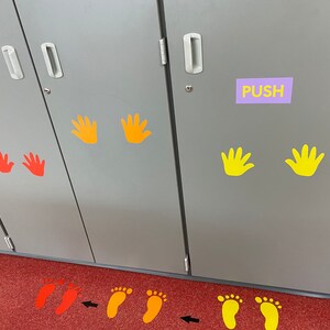 Op de afbeelding: Een grijze kast met hand- en voetgrafieken in geel, oranje en rood. Het woord "PUSH" staat op een paars label. Pijlen en voetafdrukken staan op de rode vloer en geven een pad aan.