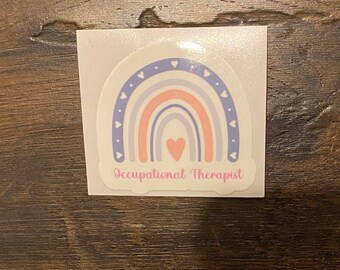 May include: A sticker with a rainbow design in shades of pink, purple, and blue. The rainbow has white hearts and blue dots along the edges. In the center of the rainbow is a pink heart. The text "Occupational Therapist" is written below the rainbow.