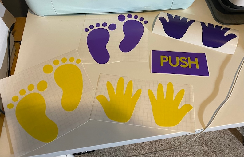 Op de afbeelding: Een verzameling vinylstickers met gele en paarse voetafdrukken, gele handafdrukken en een paars bord met het gele woord "PUSH". De stickers liggen op een wit oppervlak.