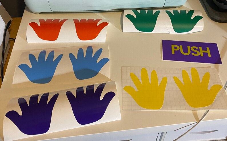 Op de afbeelding: Verschillende kleurrijke handvormige vinylstickers in rood, groen, blauw, geel en paars. Er is ook een paarse rechthoek met het woord "PUSH" in geel. Deze stickers zijn waarschijnlijk voor decoratie of bewegwijzering.