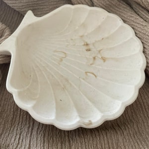 Könnte beinhalten: Eine weiße, keramik-gefertigte Schale in Muschelform mit einer strukturierten Oberfläche. Die Schale hat eine leichte braune Verfärbung.